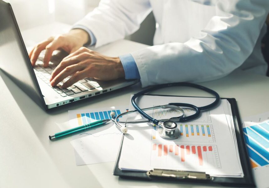 Doctor typing on laptop with stethoscope nearby.